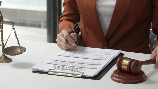 Young attractive Asian female lawyer in formal suit works on tablet with laptop, legal books, and justice scale on her desk.
