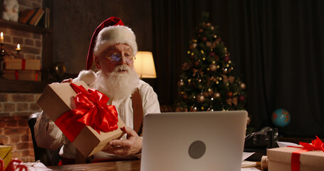 Bearded Santa Claus using laptop computer, talking on video call, holding New Year present for...