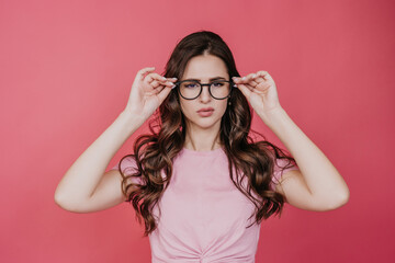 Puzzled brunette girl with wavy hair puts on glasses trying to focus looks in disbelief, standing against pink studio background.