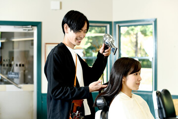 女性客の髪をドライヤーで乾かすアジア人の男性美容師 - obrazy, fototapety, plakaty