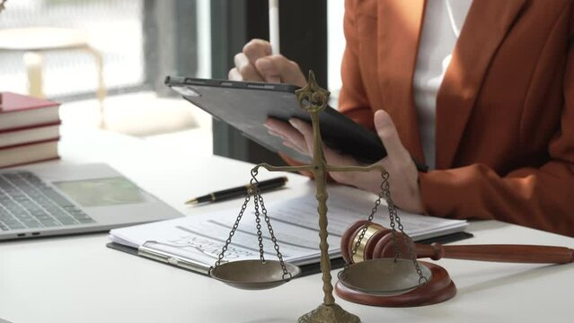 Young attractive Asian female lawyer in formal suit works on tablet with laptop, legal books, and justice scale on her desk.