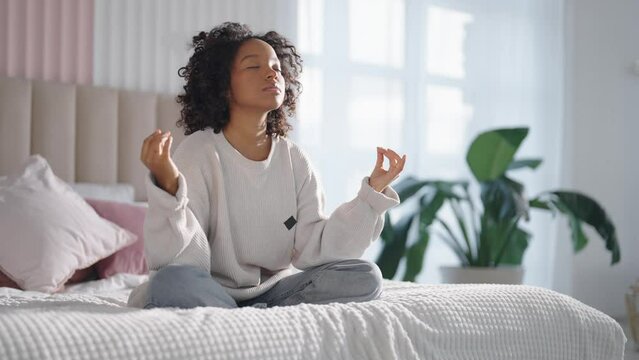 Smiling African american curly woman meditating sitting in bed at home in lotus pose feeling harmony. Girl closed eyes relaxing on meditation with hands in Gyan mudra pose. Spiritual mental practice.