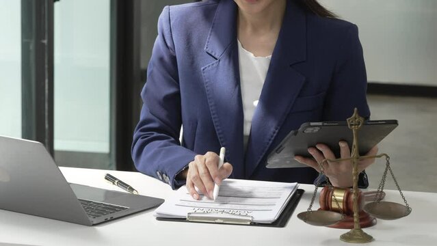 Young attractive Asian female lawyer in formal suit works on tablet with laptop, legal books, and justice scale on her desk.