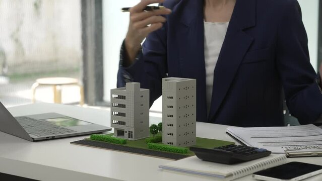 Smiling Asian businesswoman presenting model buildings, symbolizing urban investment opportunities in a city, condo, apartment.