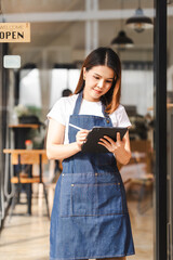 Happy excited attractive young Asian woman in denim apron, received online order in coffee shop with an 'Open' sign.