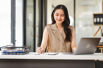 A focused Asian freelance writer takes notes at her desk with a laptop in a bright, modern office setting, journalist, artist, portrait, Thai's people.