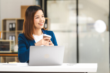 Happy attractive middle aged Asian people advertising manager business woman in formal suit smiles while using laptop, tablet, mobile phone, with laptop and coffee on desk.