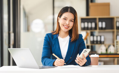 Looking camera, Happy attractive middle aged Asian people advertising manager business woman in formal suit smiles while using laptop, tablet, mobile phone, with laptop and coffee on desk.