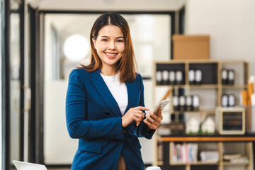 Happy attractive middle aged Asian people advertising manager business woman in formal suit smiles while using laptop, tablet, mobile phone, with laptop and coffee on desk.