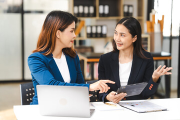 Two middle aged and young Asian female executives in formal suits review bar chart, discussing business strategies in office setting, senior executives or directors in advertising or public relations
