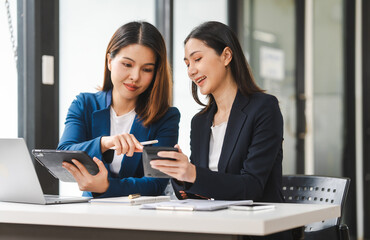 Two middle aged and young Asian female executives in formal suits review bar chart, discussing business strategies in office setting, senior executives or directors in advertising or public relations