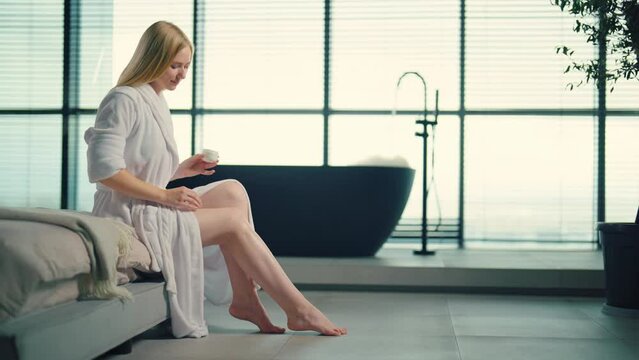 Woman applying cream on legs after bath. Wellbeing positive blonde girl enjoying doing anti-aging skincare cosmetics lifting bodycare daily treatment. Home cosmetology spa procedure self care concept.