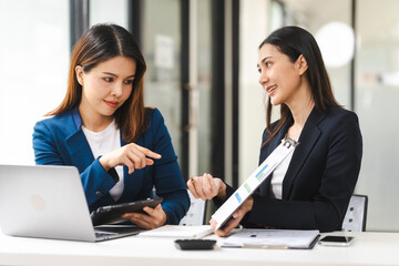 Two middle aged and young Asian female executives in formal suits review bar chart, discussing business strategies in office setting, senior executives or directors in advertising or public relations