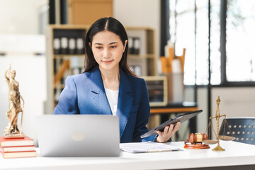 Young attractive Asian female lawyer in formal suit works on tablet with laptop, legal books, and justice scale on her desk.