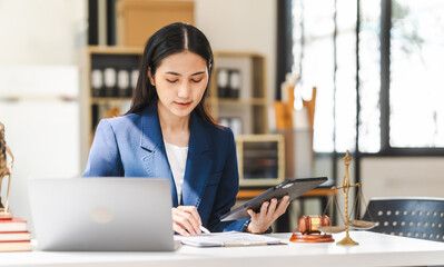 Young attractive Asian female lawyer in formal suit works on tablet with laptop, legal books, and...