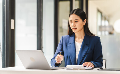 Young attractive Asian female lawyer in formal suit works on tablet with laptop, legal books, and justice scale on her desk.