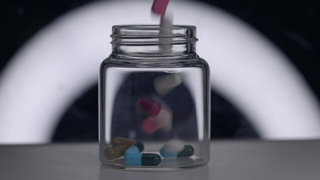 Many Pharna pills falls into glass jar filmed in slow motion - with a white half circular light in the backdrop