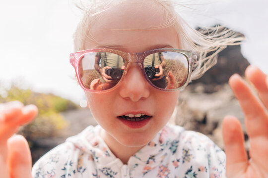 A child's curious eyes reflected in the lenses of a pair of pink sunglasses.