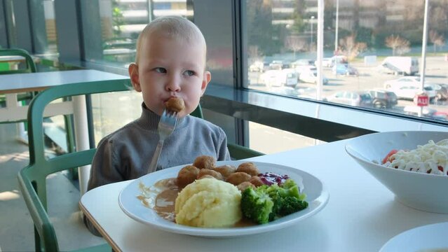 A little girl eats a meatball and mashed potatoes in a cafe near the large window of a shopping center during the day.