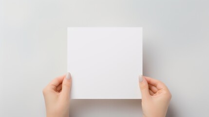 Blank white square greeting card opened by female hand with manicured nails. Mockup. Top view. Stylish and blurry background. For the text entry area - obrazy, fototapety, plakaty