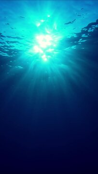 Vertical video, The sun's rays penetrate underwater into blue water through the sea waves. Underwater sunbeams, bottom view, slow motion. Underwater sun rays in the blue Ocean