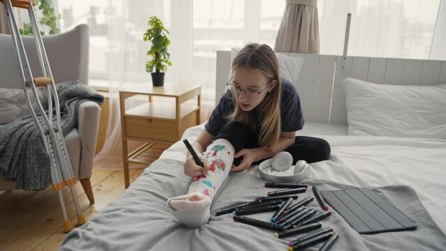 Teenage girl after injury with broken leg in orthopedic cast painting pictures felt tips sitting in bed at home. Teen in glasses drawing on cast. Recovery of damaged leg, forced sick leave concept.