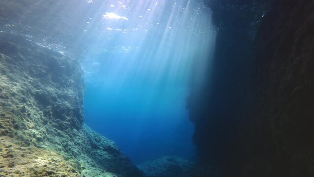 Bright dance sunrays penetrate blue water into canyon of rocky reefs, Slow motion, Camera moving forwards between cliffs in sunbeams, Underwater landscape of Mediterranean Sea