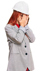 Young redhead woman wearing architect hardhat with sad expression covering face with hands while crying. depression concept.