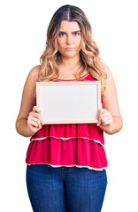 Fototapeta premium Young caucasian woman holding empty white chalkboard thinking attitude and sober expression looking self confident