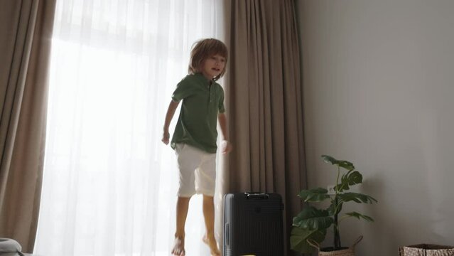 Cute happy child, fair-haired boy of 6-7 years old, is joyfully jumping on overfilled suitcase with things sticking out of it. Concept of adventure and anticipation. Advertisement of app for trip 