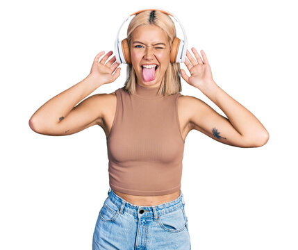 Beautiful young blonde woman listening to music using headphones sticking tongue out happy with funny expression.