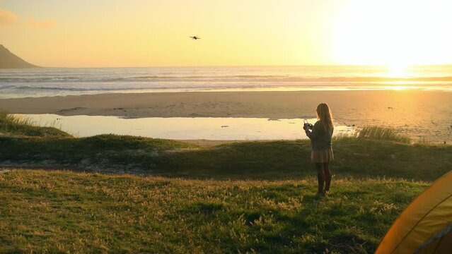 Woman pilot drone at beach, sunset and quadcopter flight at sea, spy or surveillance on summer holiday travel. UAV, back and helicopter in air, future robot tech and photography for camping at tent