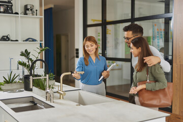 Female shop assistant helping young couple choose new kitchen faucet