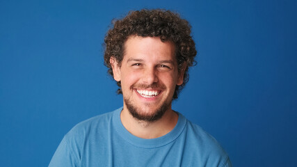 Close up, guy with curly hair dressed in blue t-shirt looks at the camera with smile isolated on...