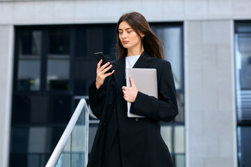 Powerful woman multitasking with her phone and laptop while standing outside among towering office buildings