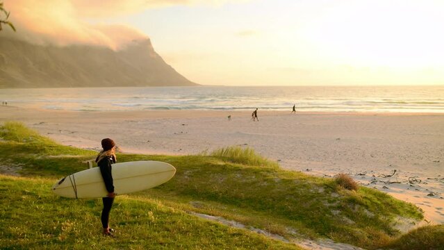 Sunset, freedom and woman with surfboard on beach with mountain, cloudy sky and view of ocean on holiday. Adventure, travel and girl on tropical vacation for surfing, peace and calm sea on island.