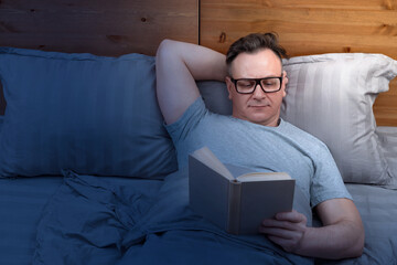 A man with glasses is lying on the bed reading a book before going to bed, by the light of a night...