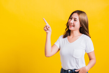 Asian Thai happy portrait beautiful cute young woman standing wear white t-shirt pointing finger away side looking to away side, studio shot isolated on yellow background with copy space - obrazy, fototapety, plakaty