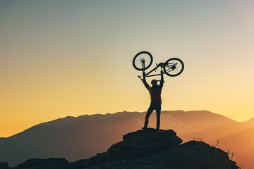 Happy biker stands in winner success pose with mtb bike above his head at mountain top and looks at...