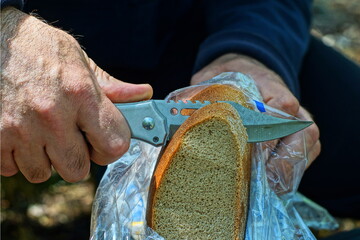 a man with an iron sharp folding pocket army knife in his hand cuts a piece of fresh tasty gray bread from a round loaf on the street during a picnic during the day - obrazy, fototapety, plakaty