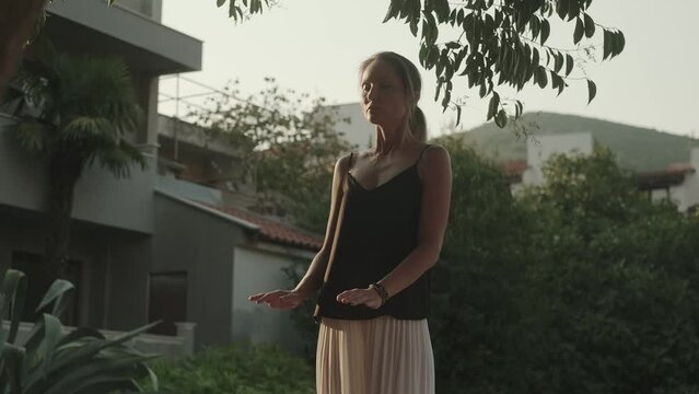 One white woman in a black sleeveless tank top practices slow controlled movements with her eyes closed outdoors. Asian meditation exercises. Qigong. T'ai chi practice. Chinese psychopractice.