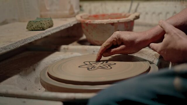 The artist draws a clay flower on spinning pottery wheel. Craftsman workshop atmosphere. Masterclass of drawing plate ornament at dirty workplace. Drawing skills