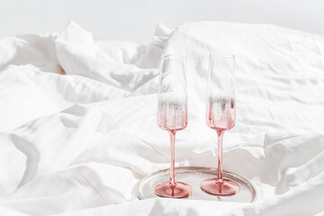 Two pink colored shiny champagne glasses with rose wine on bed, on white sheets. Minimal lifestyle...