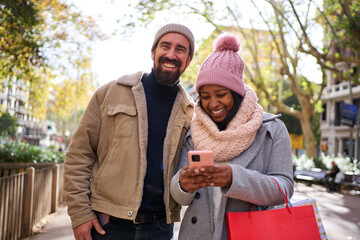 Happy interracial young couple doing christmas shopping in the city, standing outdoors using smart...