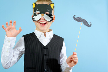 An elegant boy in a suit and carnival cat mask and holding mustache on a stick on the blue...