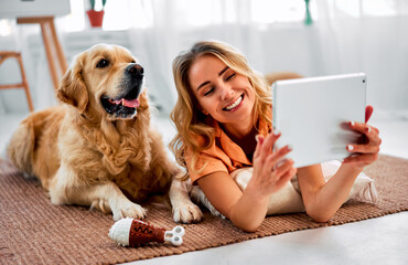 Video call with family. Communication with an animal. A happy beautiful woman is lying in the living room next to a fluffy dog and holding a digital tablet.