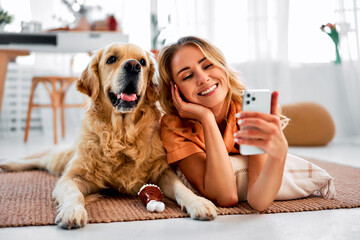 Saving memories with pet. Smiling woman with blond hair snuggling to furry friend and taking selfie on modern cell phone. Obedient golden retriever lying on floor near delighted female owner. - obrazy, fototapety, plakaty