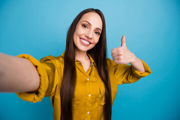 Photo of positive toothy woman dressed yellow shirt tacking selfie showing thumb up isolated blue...
