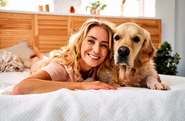 Tranquil moment at home. Fluffy family dog resting on comfy white bed with charming female owner. Woman smiling sincerely smiling at camera while cuddling with lovely labrador.