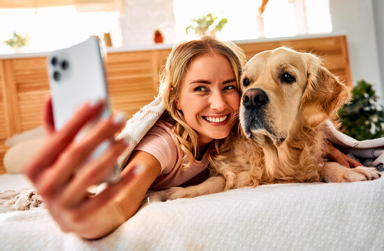 Relaxed home setting. Adorable beautiful woman using modern smartphone for taking selfie with golden retriever in bed. Smiling female embracing favorite pet and enjoying positive moment.